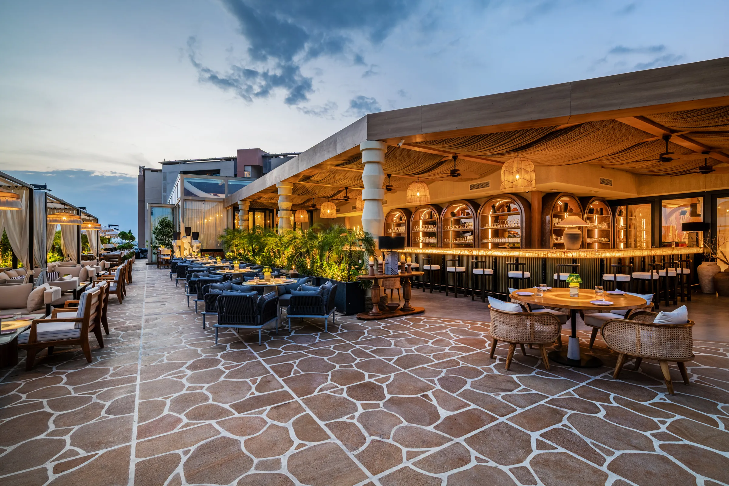 The open-air terrace at dusk — cane chairs on crazy-paving stone, the bar glowing under the rafters
